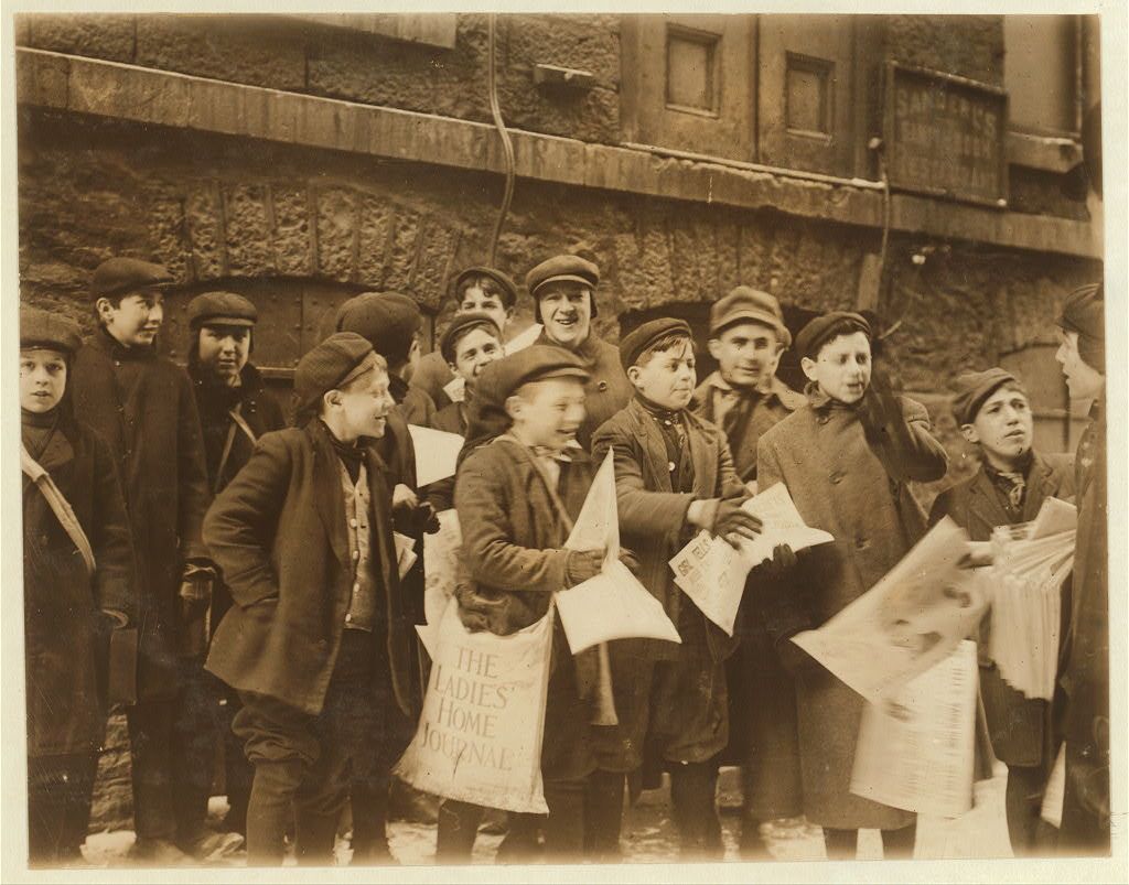 A group of cheerful-looking young boys in wool coats and hats are caught in motion as they stand to receive their loads of newspapers for sale.