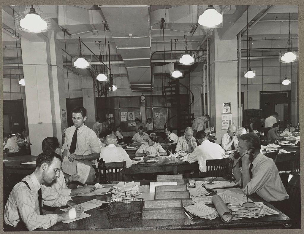 A black and white photo of a midcentury newsroom. A small platoon of young-to-middle aged white men in wrinkled shirts sit at paper-strewn desks tethered to the ground by old-fashioned telphones.
