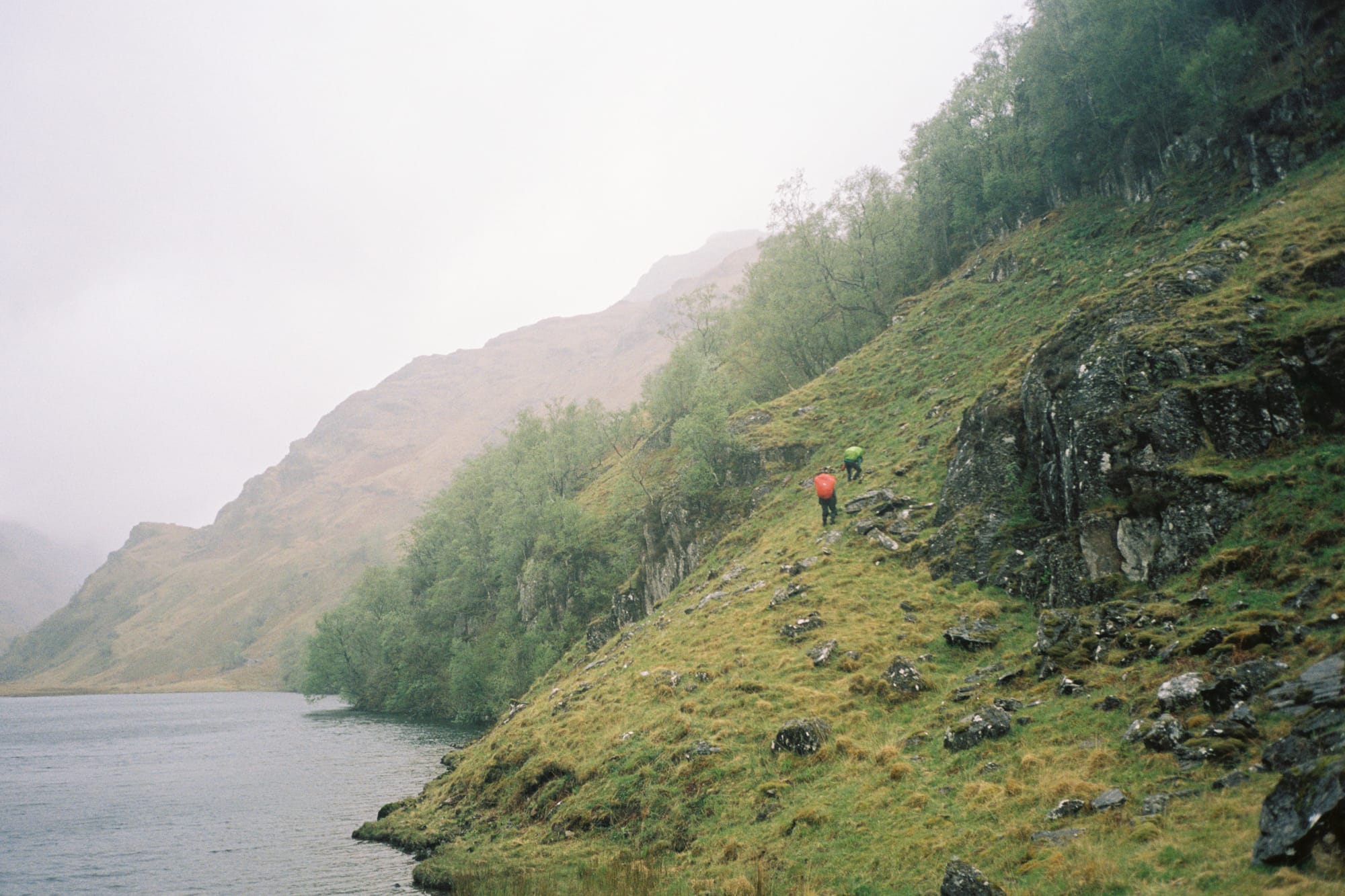 A photo of a hike in Knoydart, Scotland. Two people are in the distance hiking up a munro on the coast near a body of water.