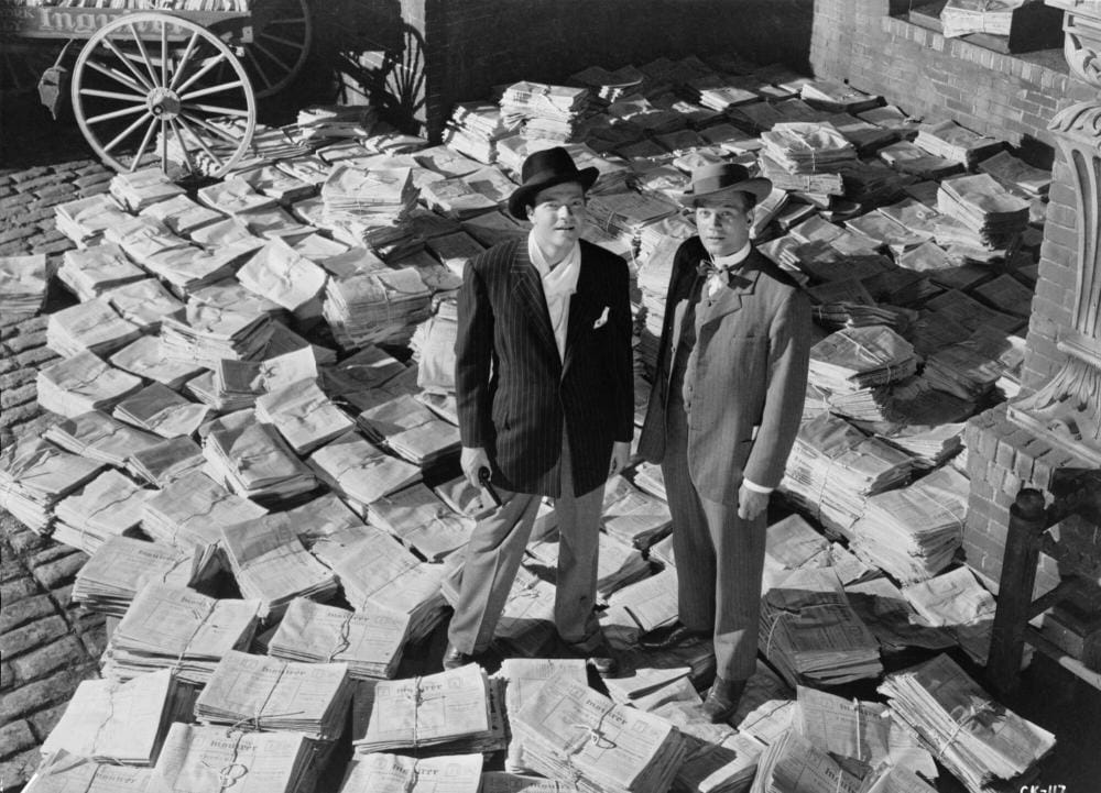 Two men in dandyish suits stand athwart a colossal pile of bound newspapers.