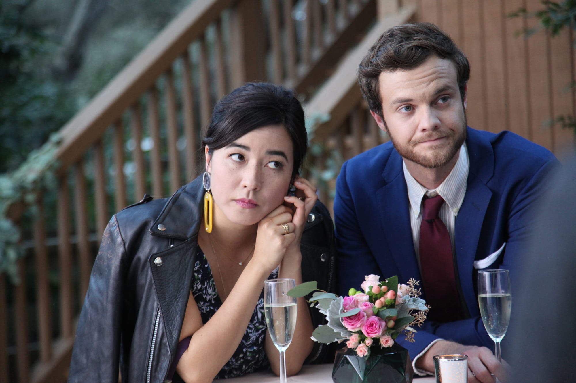 Maya Erskine and Jack Quaid sit looking unimpressed with their glasses of champagne at a reception.