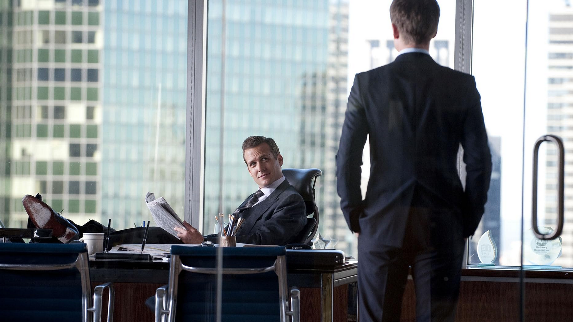 A smirking Gabriel Macht regards Patrick J Adams with the New York Skyline behind him.