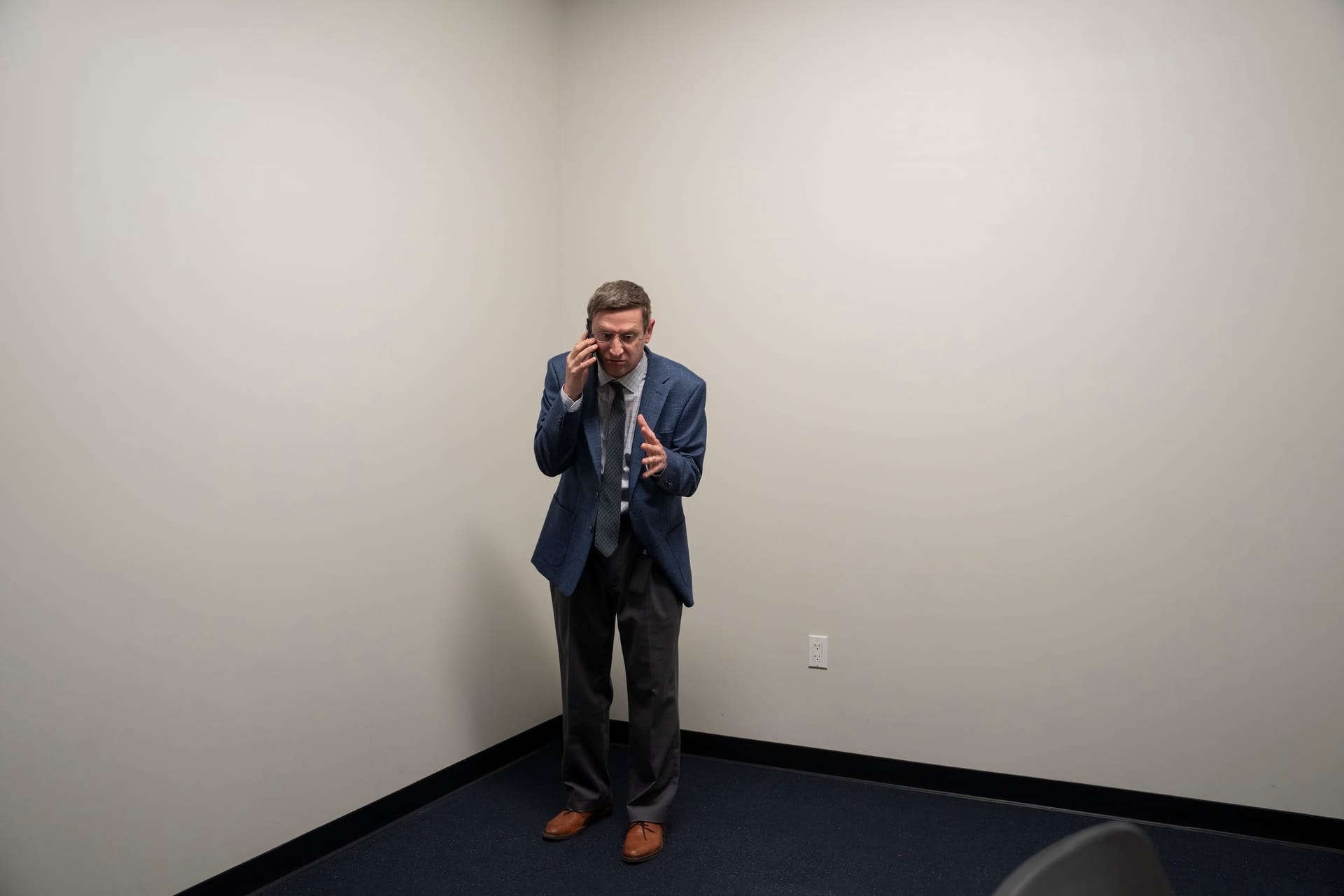 Tim Robinson wearing a blue suit and standing in the corner of a bland, white walled room, looks frustrated as he speaks on the phone.